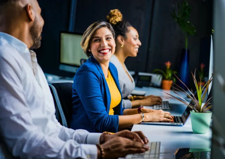Illustrative photo: three colleagues chat at their desks with a tasteful office plant and neat office fashion
