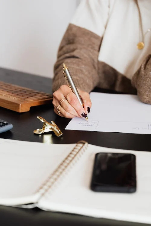 Illustrative photo: close-up of someone handwriting on paper with a keyboard and phone on the desk.