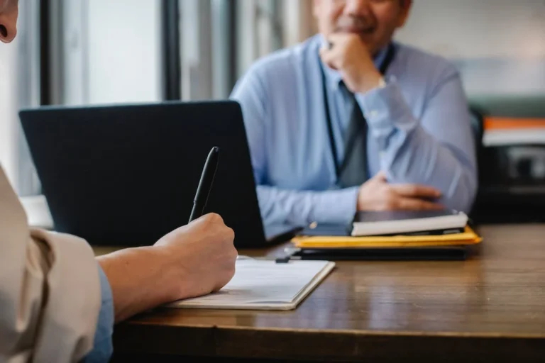 Illustration photo: two people holding a meeting across a table with one writing and presumably talking, while the other is listening