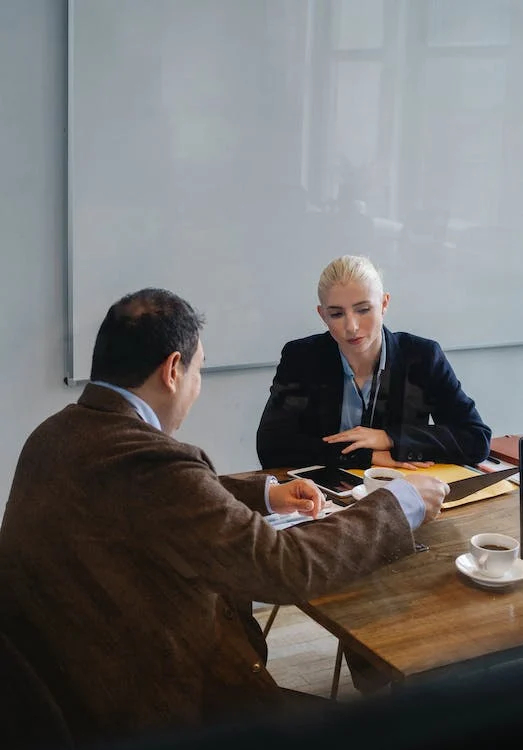 Illustration photo: two business people sit at the corner of a table, one showing the other a screen or document